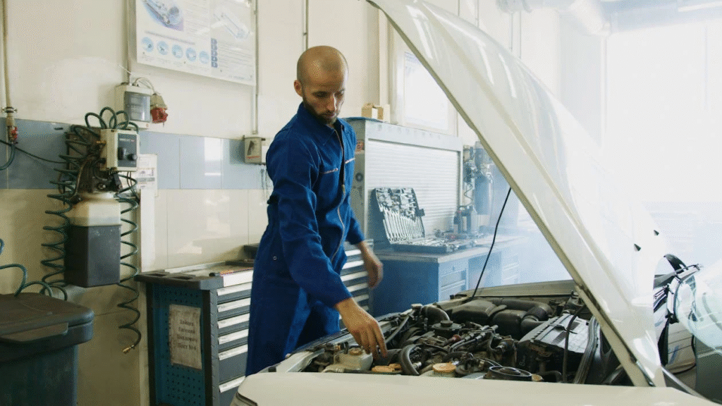 A man in blue overalls repairs a car, focused on the engine under the hood in a garage setting.